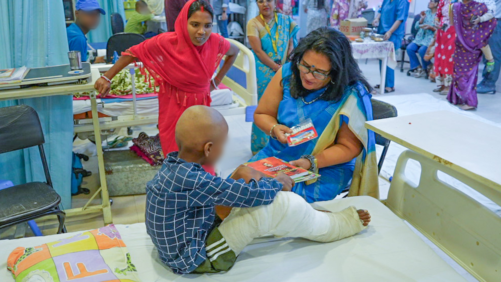 Poonam Bagai, a woman with a warm smile, sitting closely and comforting a young child wearing a medical mask in a brightly colored hospital setting